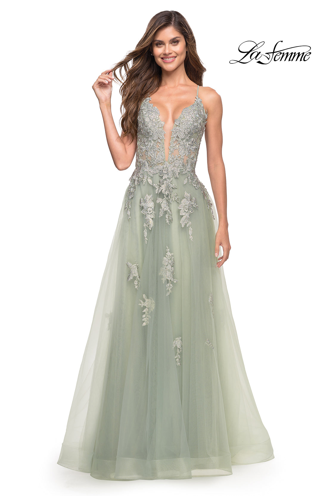 A woman smiles while standing in a sage green La Femme Open-Back Long Ball Gown with lace, featuring a plunging neckline and silver floral embroidery.
